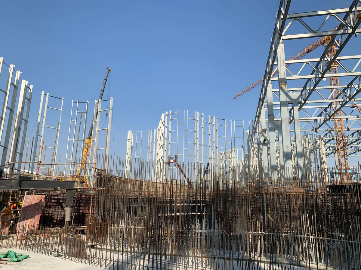 Steel framework being erected at a construction site with cranes under a clear blue sky.