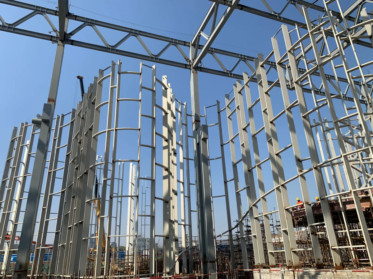 Steel building frame under construction with tall vertical columns and cross bracing; workers on a platform amid the framework.