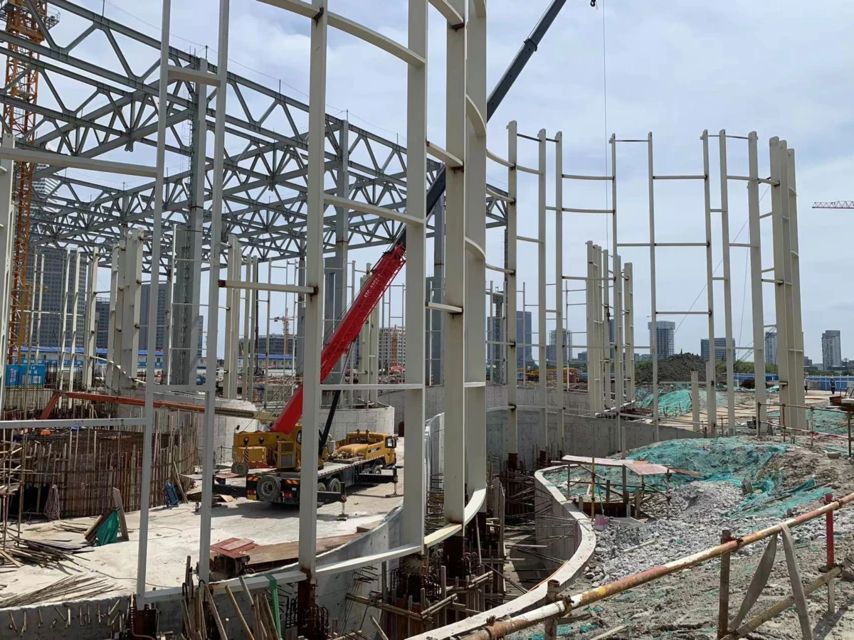 Steel-framed construction site with a red crane arm and yellow mobile crane; city skyline in the background.
