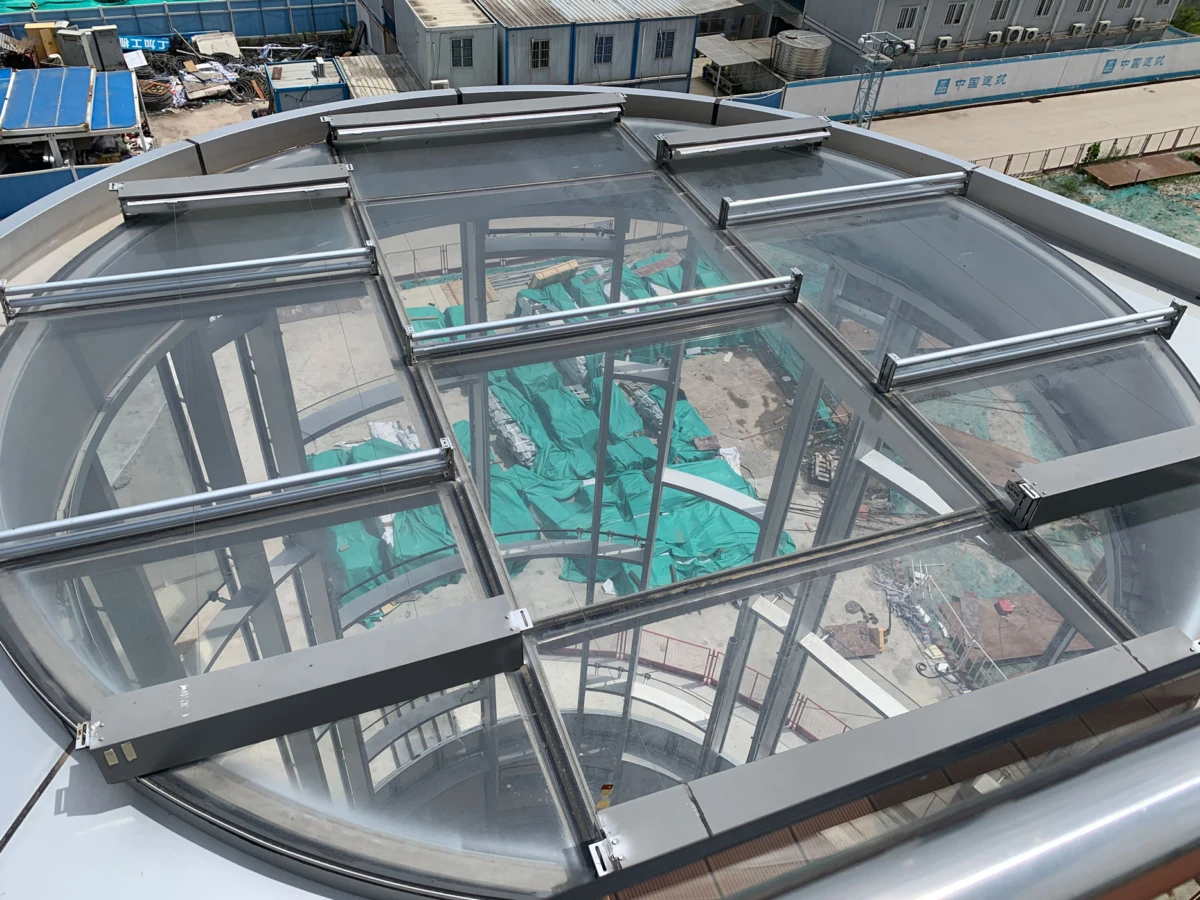 Curved glass skylight with steel framework atop a building, viewed from above; construction site with green tarps and portable buildings in the background.