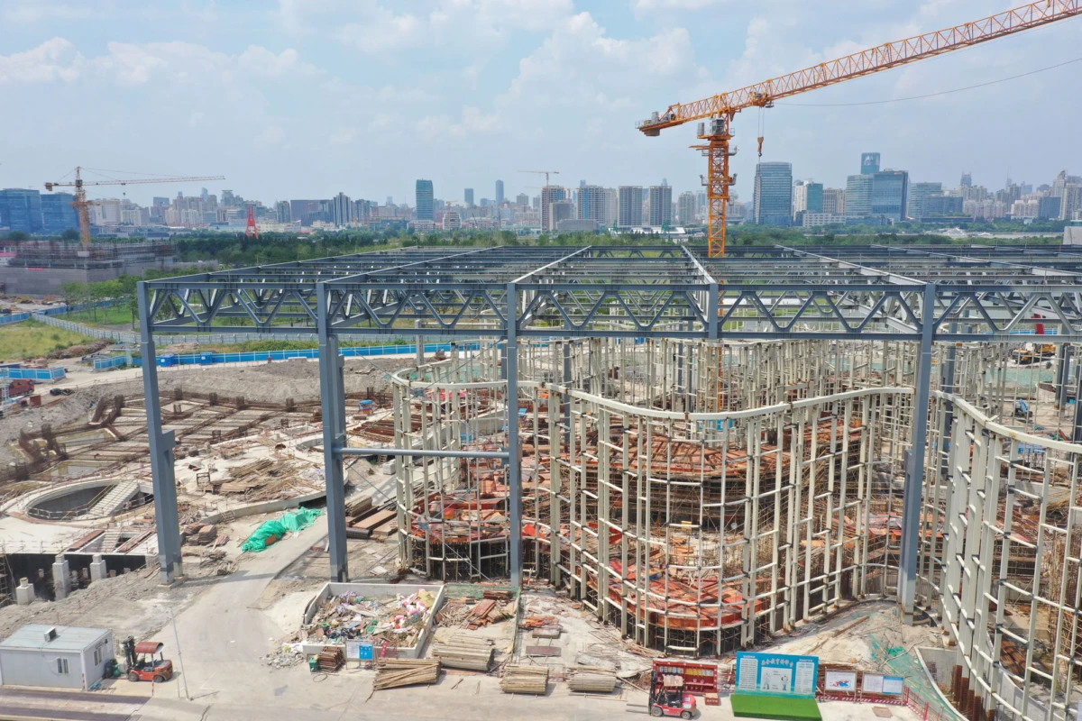 Circular concrete formwork being constructed inside a steel framework at a construction site; orange crane overhead, city skyline in distance.