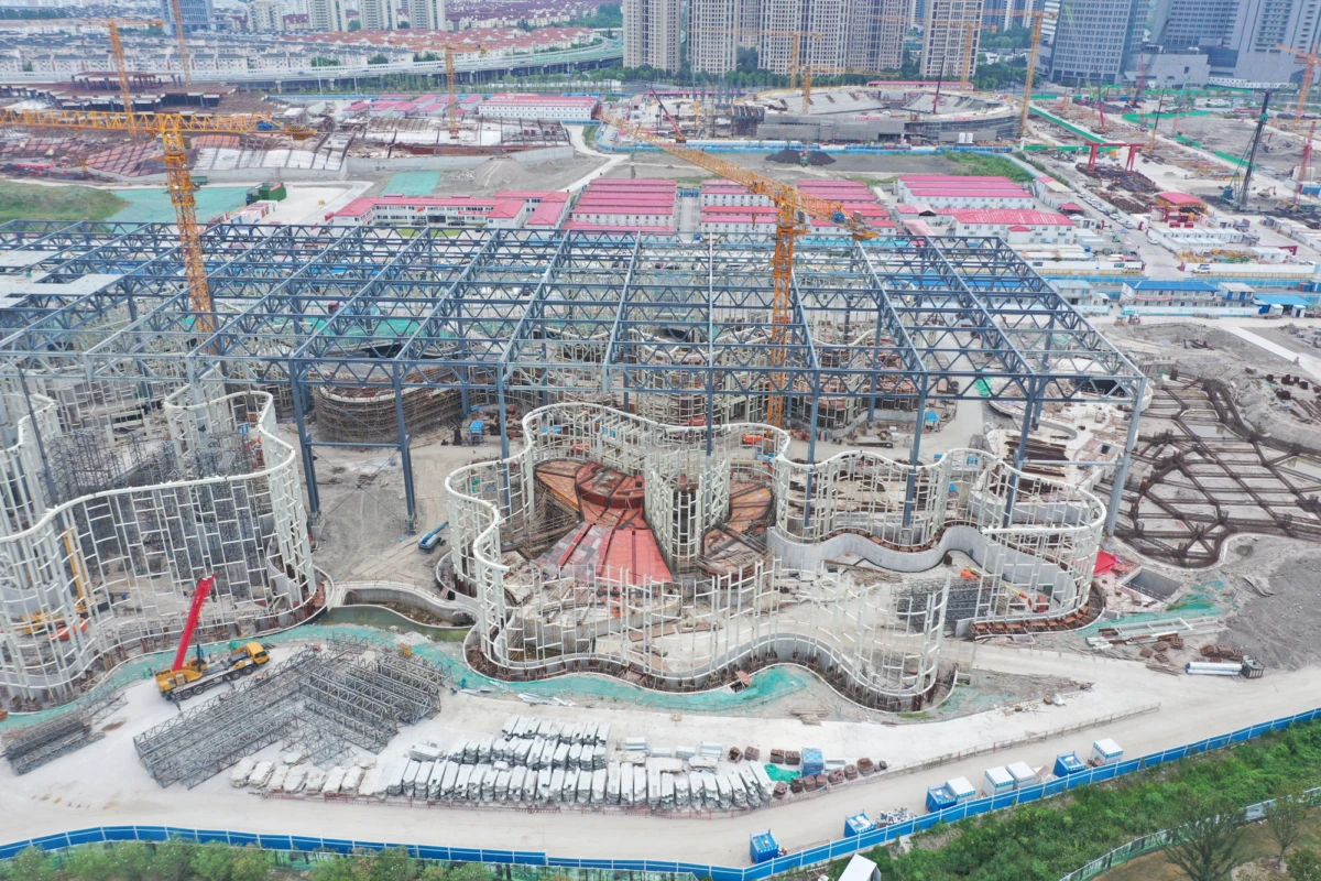 Under-construction steel-framed building with curved white facade elements; cranes and equipment on a city site with skyscrapers in background.