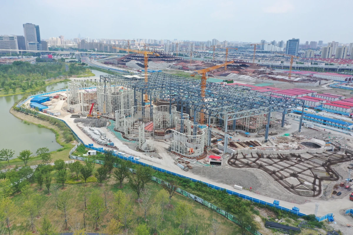 Construction site with steel framework and tall cranes; building under construction near a river, with an urban skyline in the background.