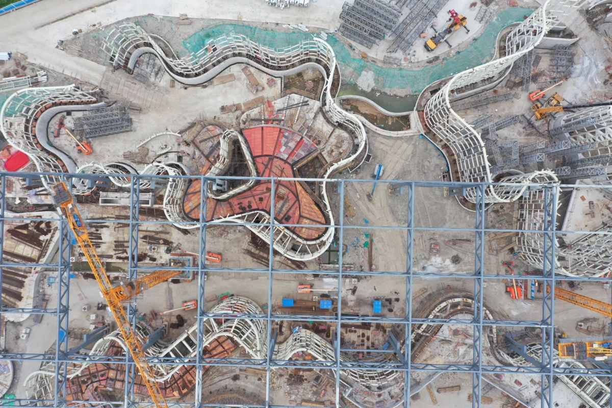 Aerial view of a construction site with sinuous white steel ramps around a red circular core, blue lattice scaffolding, and orange cranes.