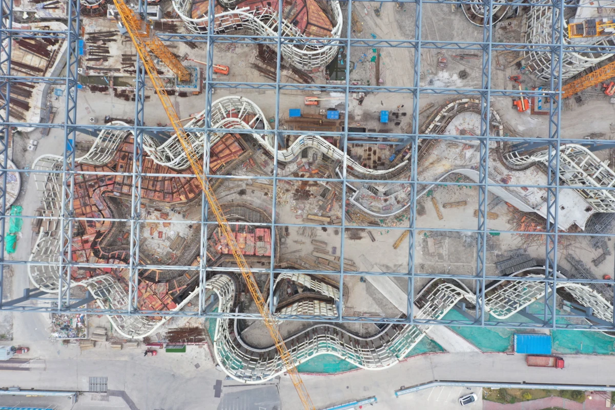 Aerial view of a construction site with curved white concrete ramps within a blue steel grid, crossed by a yellow crane.