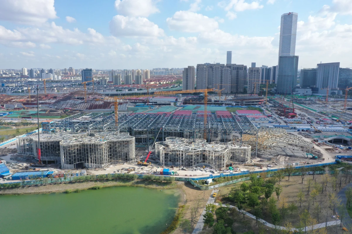 Construction site with circular steel-framed structures under construction, cranes overhead, by a river with a city skyline in the background.