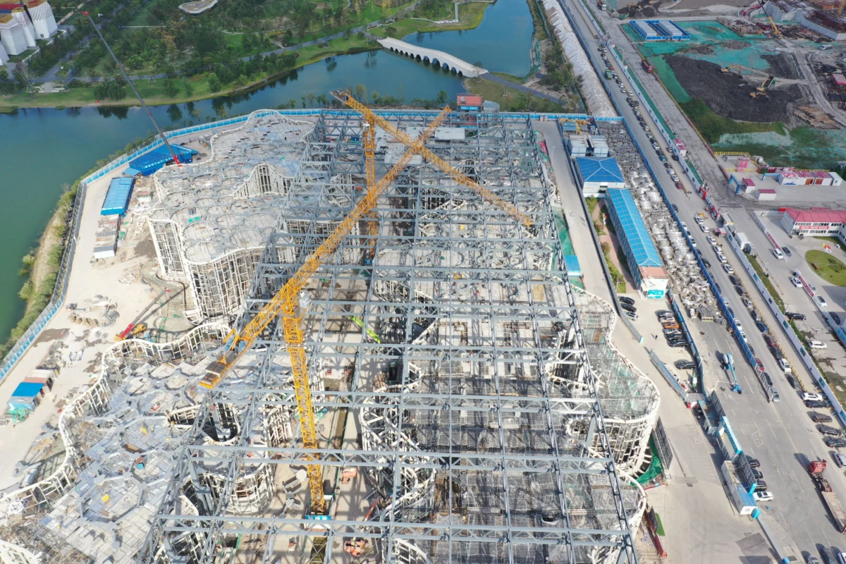 Aerial view of a large steel-framed building under construction with a yellow crane, beside a river and road.