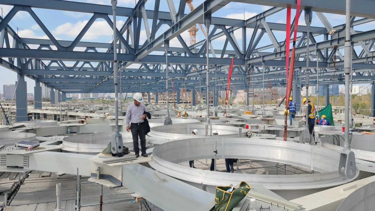 Worker in hard hat on circular metal platform inside a steel-framed industrial construction site; other crew members visible in background.