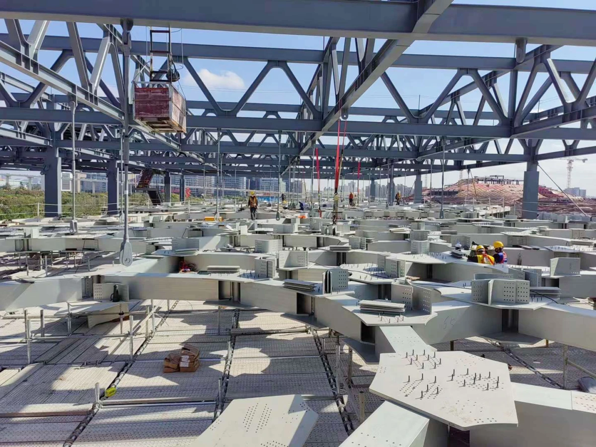 Construction site inside a large steel framework; workers install curved steel beams and platforms beneath crisscrossing girders.