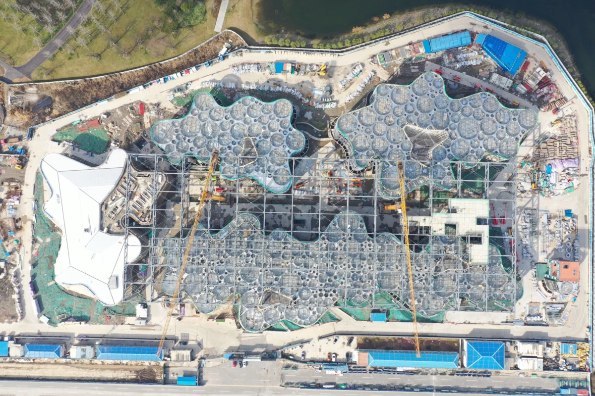 Aerial view of a construction site with a large building under construction featuring interconnected circular modular roofs; cranes on site near a river.