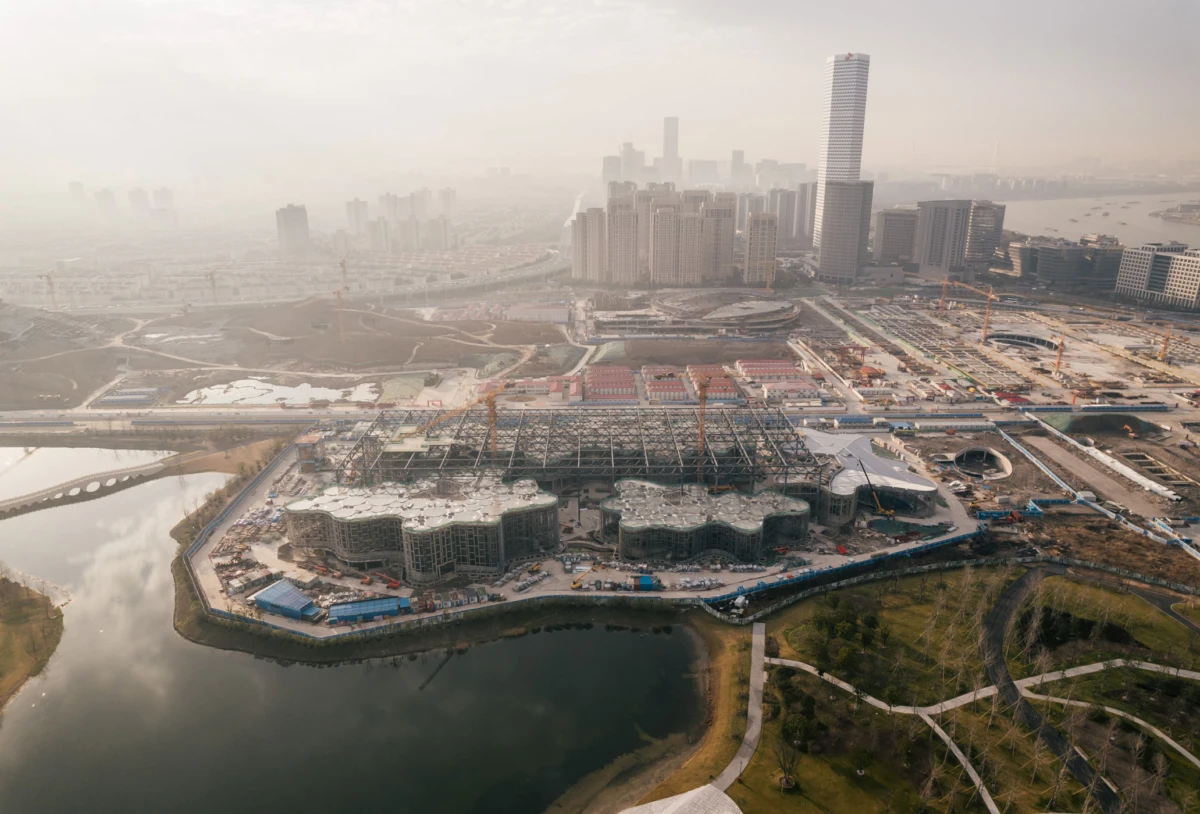 Aerial view of a large construction site with cranes beside a curved glass-building complex by a river, with a dense urban skyline.