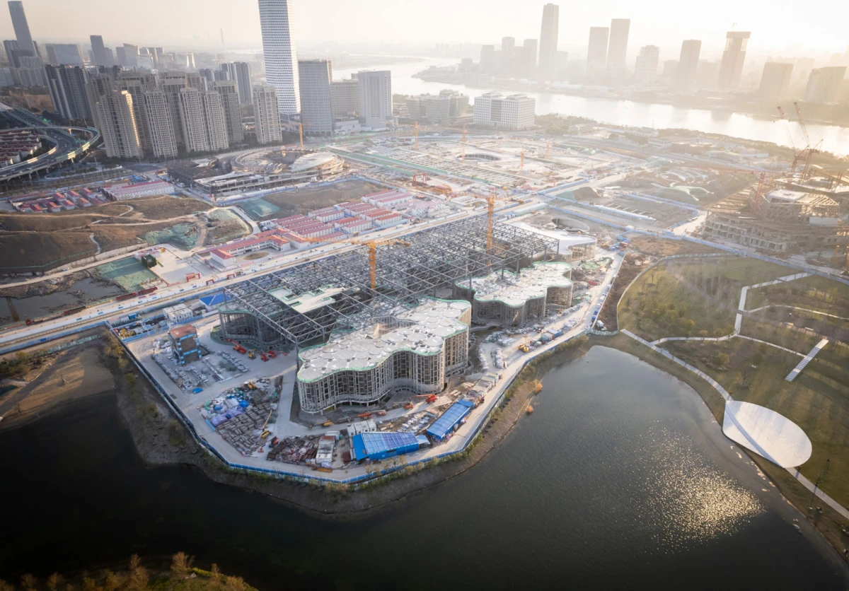 Aerial view of a large construction site with curved, glass-fronted buildings under development, cranes overhead, beside a river and urban skyline.