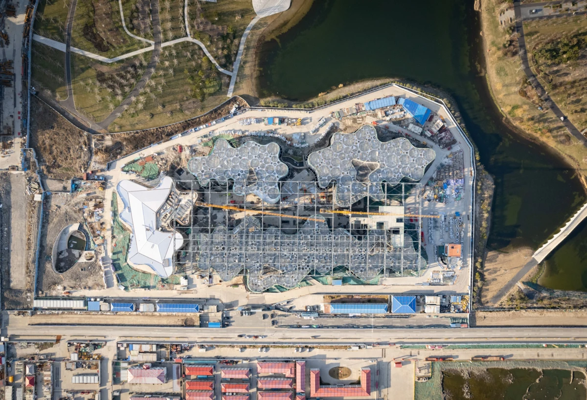 Aerial view of a construction site with a large irregular honeycomb roof structure beside a river.