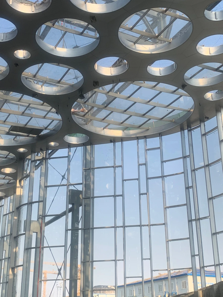 Interior of a glass-walled atrium with circular skylights in a metal ceiling, exposing steel beams and blue sky outside.