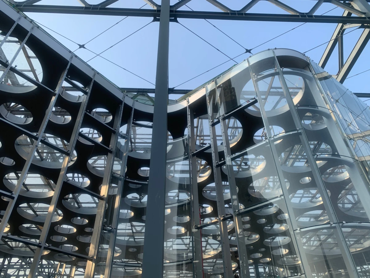 Interior of a modern glass-and-steel atrium with circular perforated metal panels and exposed beams, viewed upward.