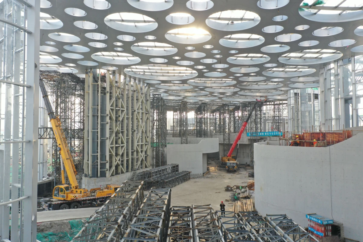 Interior of a large construction site with cranes, scaffolding, and concrete walls beneath a ceiling of circular cutouts.