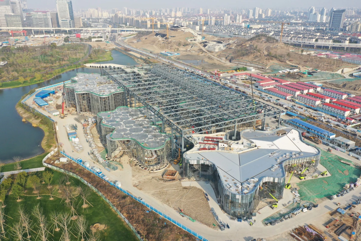 Aerial view of a sprawling construction site with interconnected curved-glass pavilions and steel framework beside a river in a developing city.