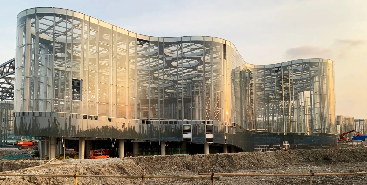 Curved glass building under construction at a dusty site, sunlight reflecting off the façade.