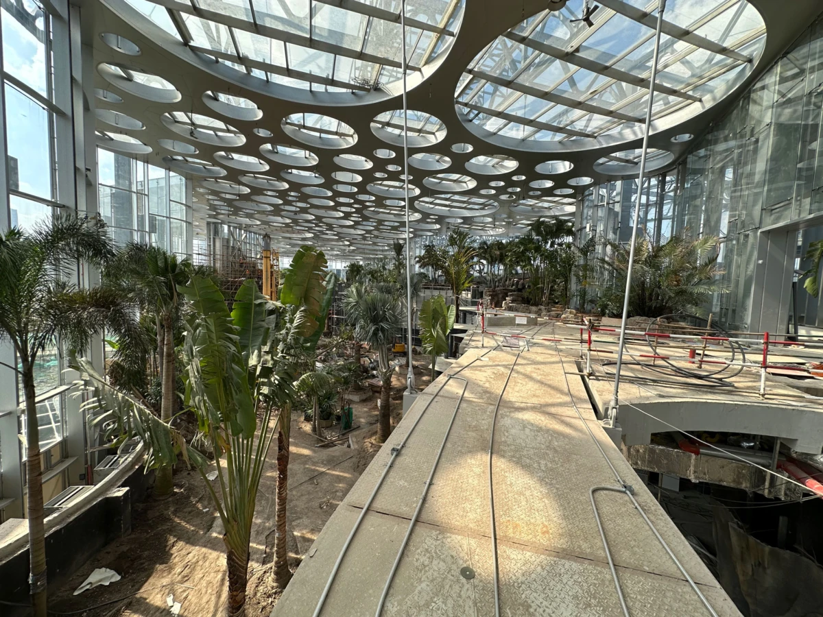 Indoor botanical conservatory with tropical plants along a raised walkway under a circular skylight ceiling.