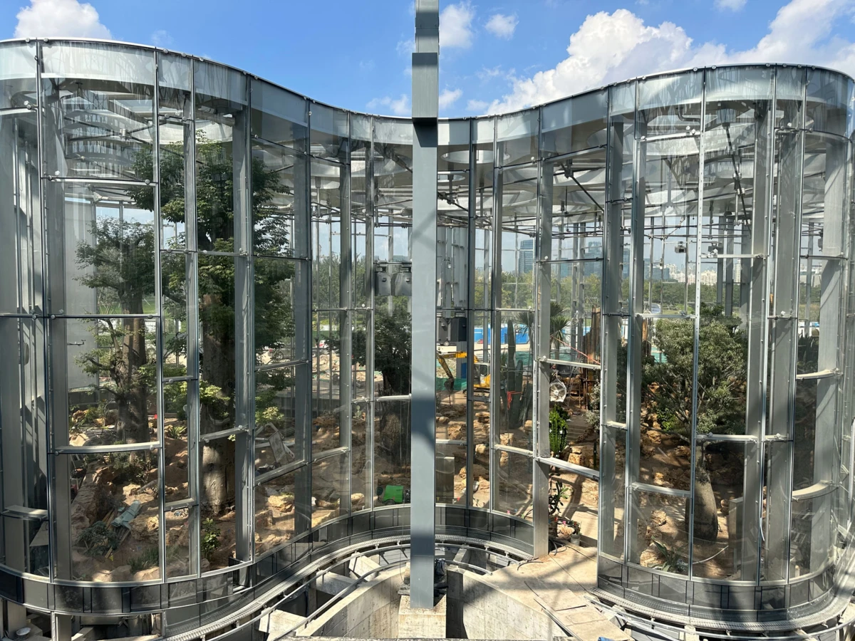 Circular glass conservatory with curved walls and metal framework, housing an indoor garden of trees under a blue sky.