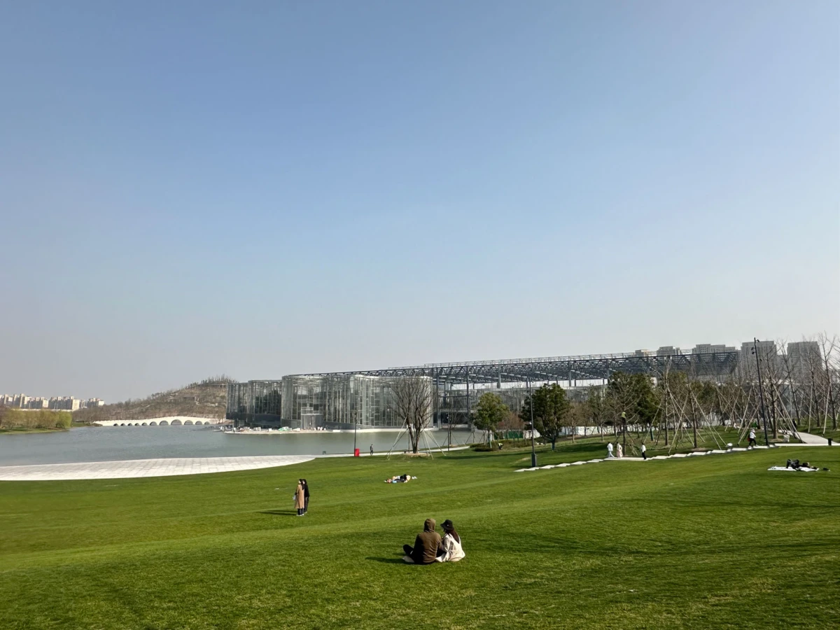 People sitting on a grassy park by a lake, with a large glass-fronted modern building in the background.