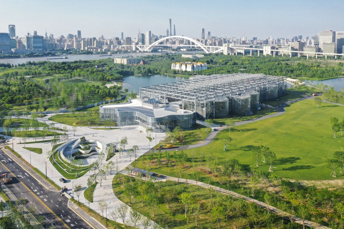 Futuristic glass-pavilion complex sits in a riverside park, with an arched bridge and city skyline in the background.