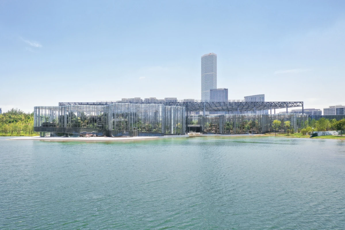 Glass-walled modern pavilion on a lake, with a metal framework, surrounded by trees; urban skyline in the distance.