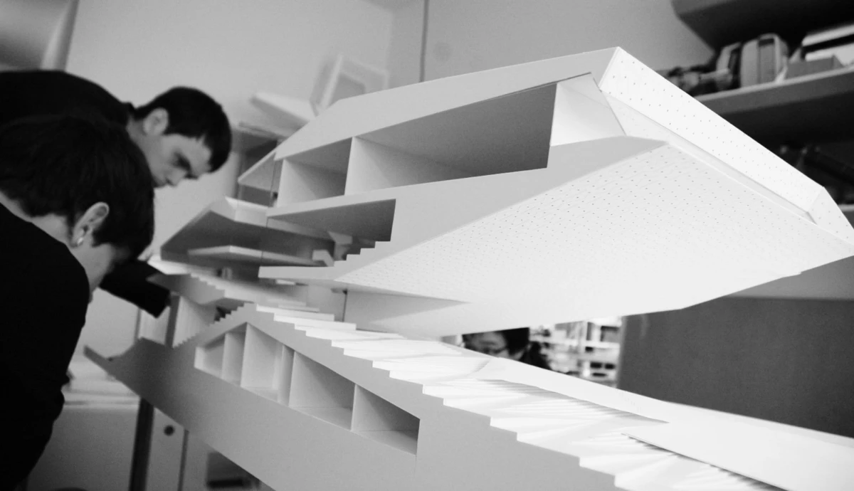 Two designers assemble a large white architectural model with stacked, stair-step panels in a studio.