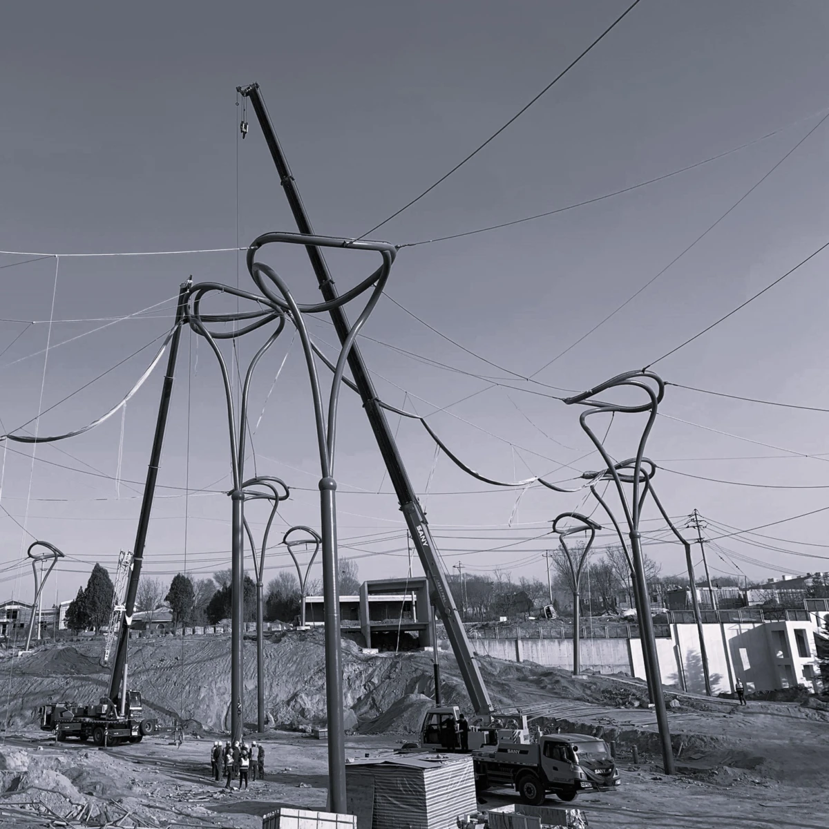 Construction site with tall looping curved steel frames; crane lifts a beam as workers stand on dirt ground.