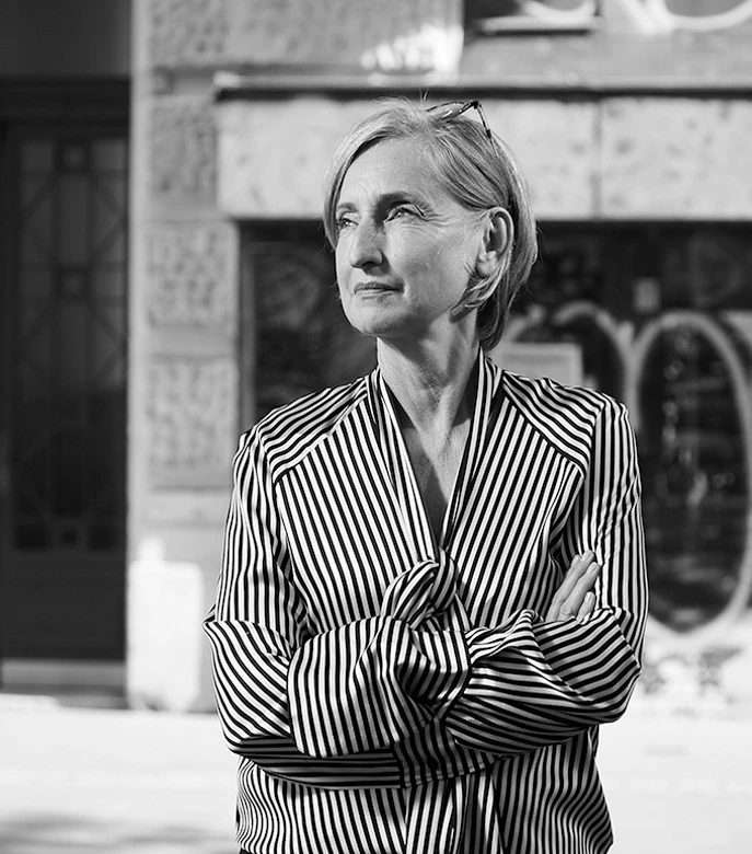 Person with short gray hair in a striped blouse, arms crossed, looking left in an urban street setting.