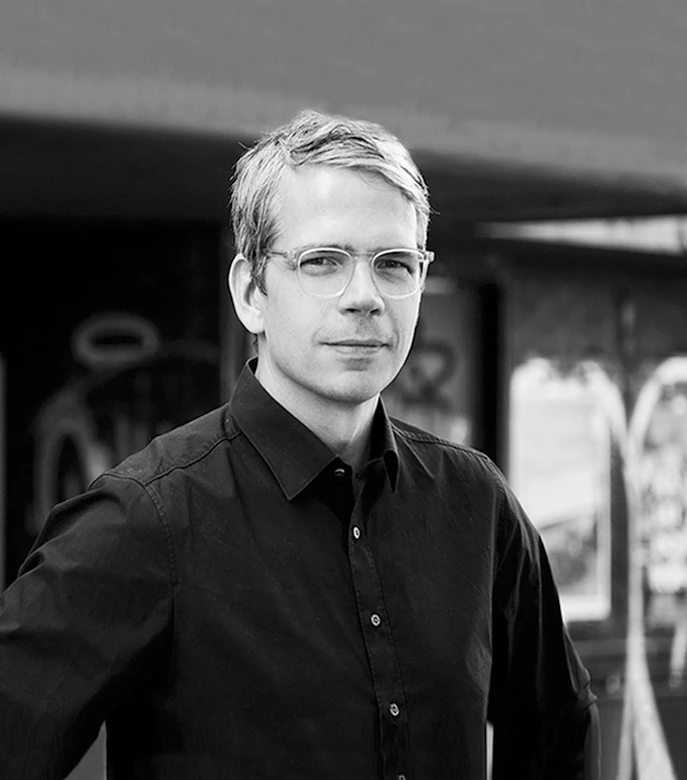 Adult person with light hair and rectangular glasses stands outdoors in an urban storefront setting, facing the camera.
