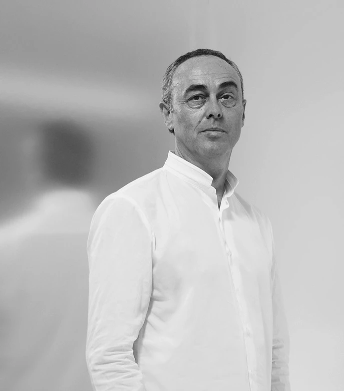 Middle-aged man in a white shirt stands in a plain indoor setting, facing the camera with a slight turn and a calm expression.