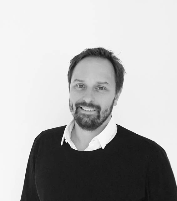 Adult with beard smiling, wearing a white collared shirt under a black sweater, standing against a white background.