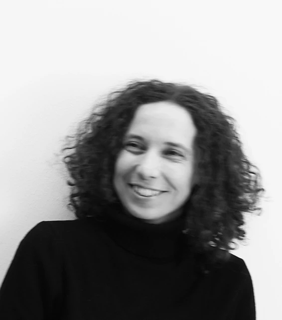 Portrait of a person with curly hair wearing a black turtleneck, smiling, against a plain light wall.