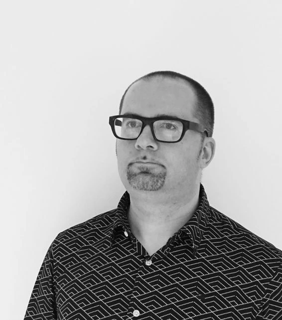 Adult person with short hair, goatee, black rectangular glasses, wearing a geometric-patterned shirt, against a plain background.