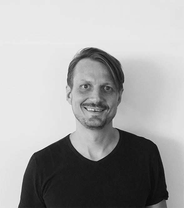 Adult person with short hair wearing a black t-shirt smiles at the camera against a plain light wall.