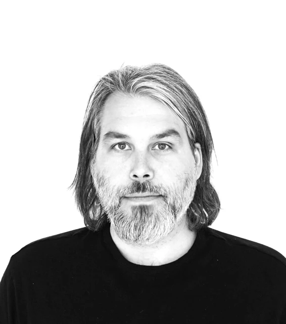Adult with shoulder-length gray hair and beard, wearing a black shirt, posing for a studio portrait against a white background.