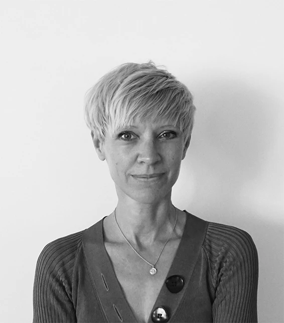Woman with short blonde pixie haircut, wearing a cardigan and pendant necklace, posing against a plain wall.