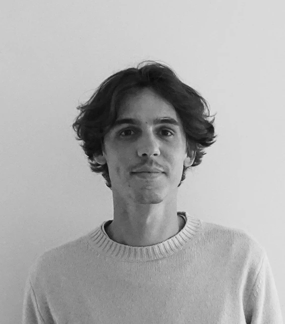 Young adult with shoulder-length wavy hair wearing a light sweater, facing the camera against a plain white wall.