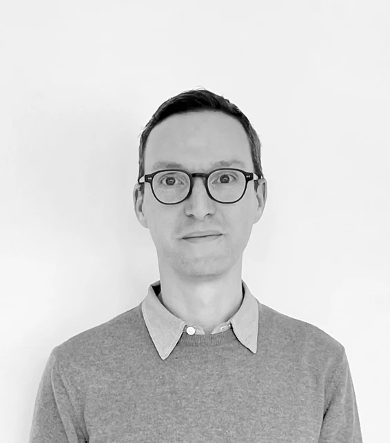 Adult person with short dark hair and rectangular glasses, wearing a gray collared sweater, standing against a white background.