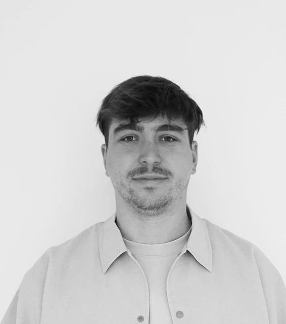 Portrait of a person with short dark hair and light stubble, facing the camera against a plain white background, wearing a light collared shirt.