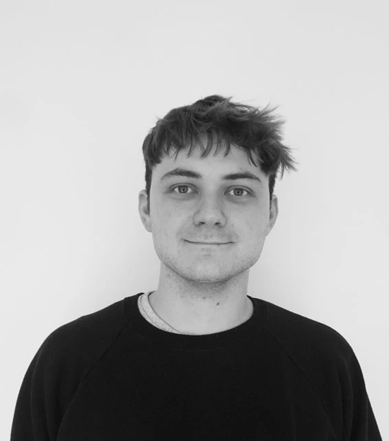 Young adult with short dark hair, facing the camera with a neutral expression, wearing a black crew-neck sweater against a plain white background.