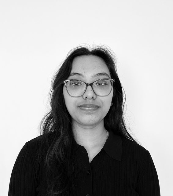 Portrait of a person with long dark hair and glasses, looking at the camera, wearing a black collared sweater against a white background.