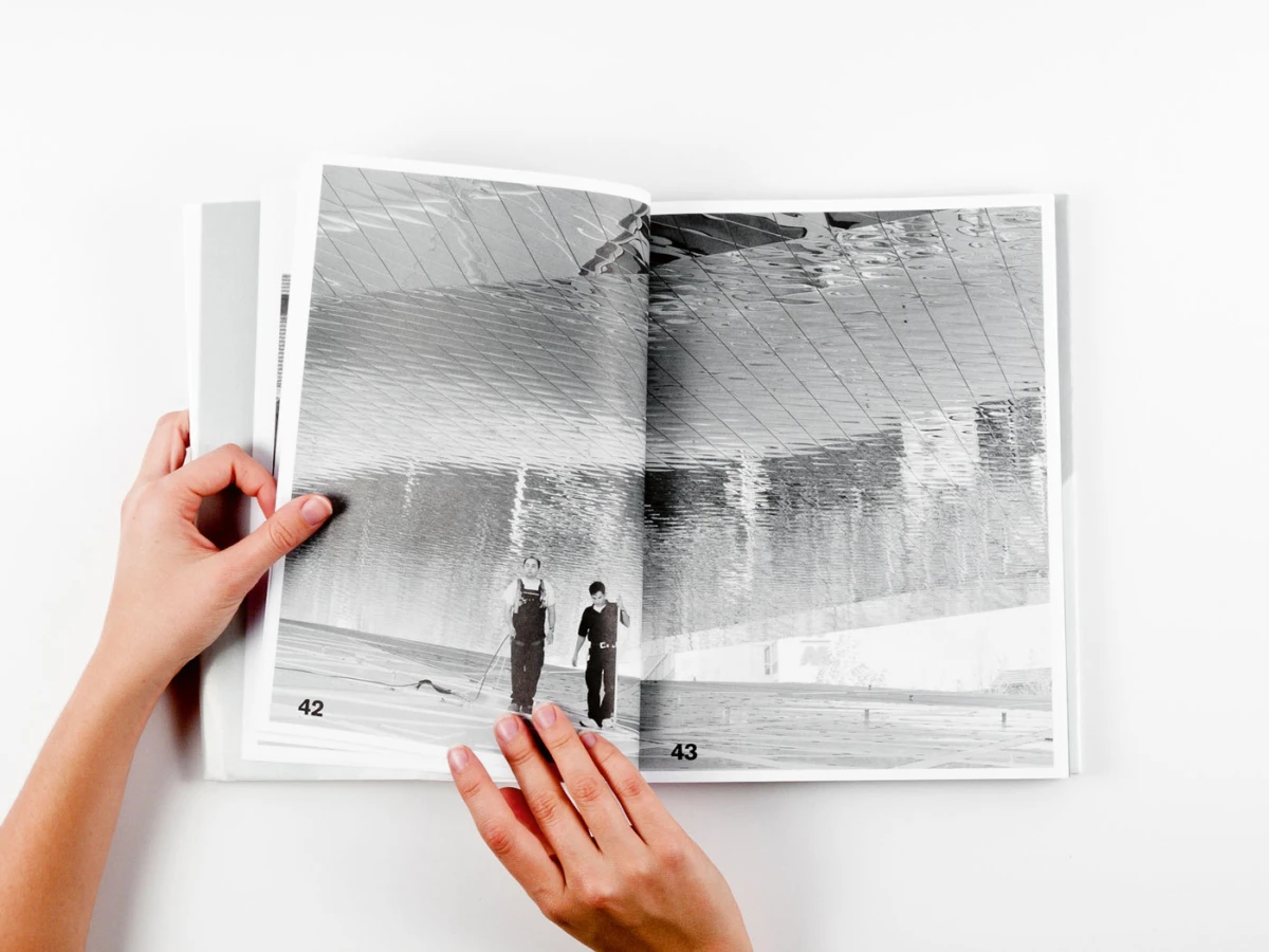 Hands turning pages of a black-and-white photo book on a white surface, revealing two people indoors.