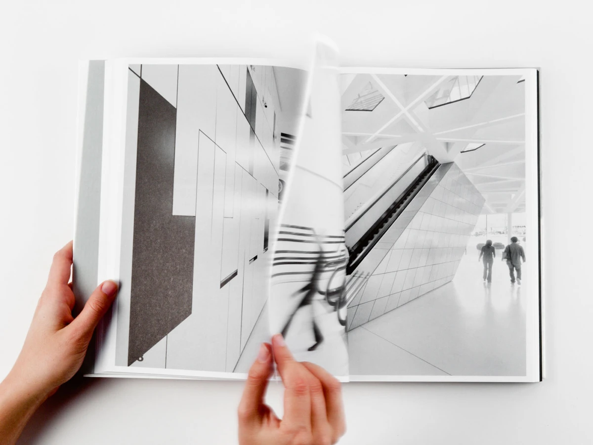 Hands turning pages of an open photobook revealing a black-and-white architectural interior with an escalator and two pedestrians.