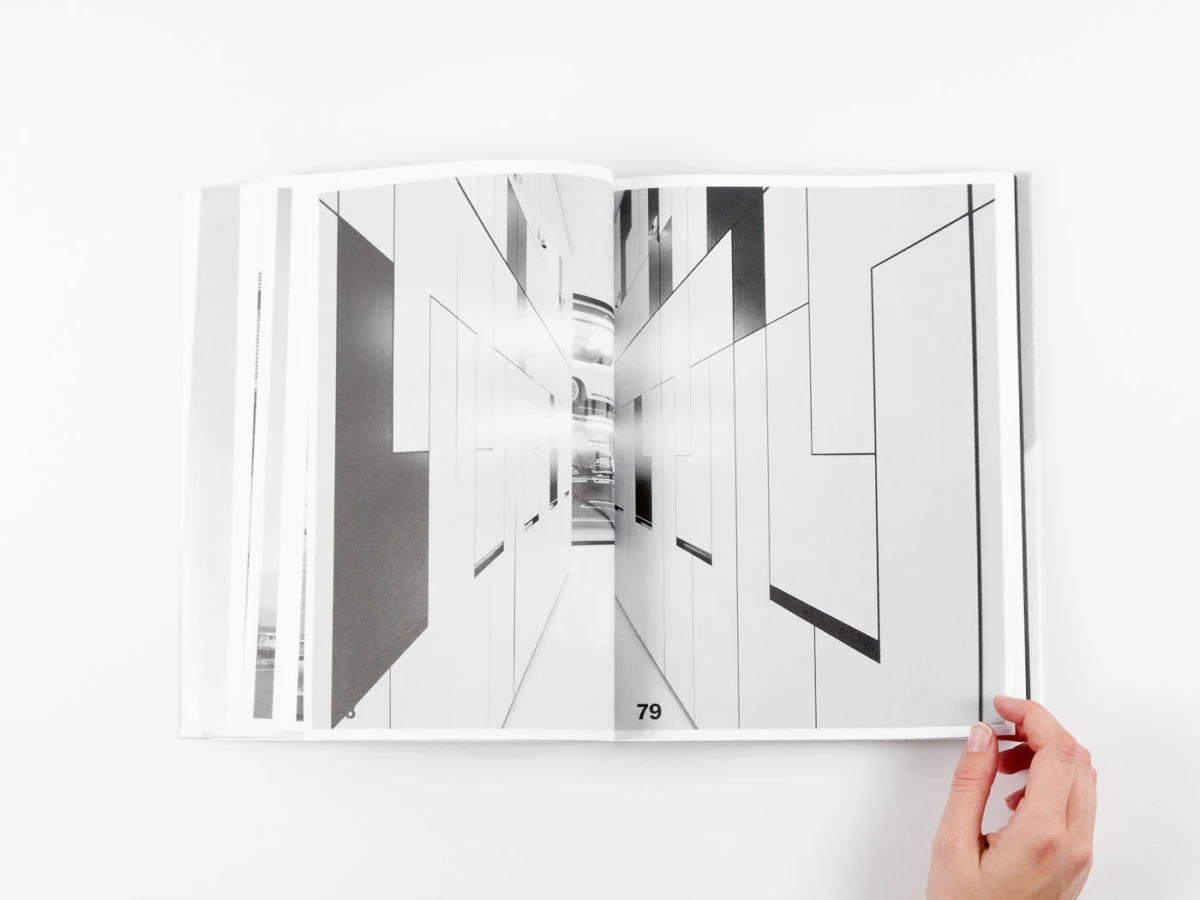 Open book on a white surface showing a black-and-white architectural corridor spread; a hand holds the right edge.