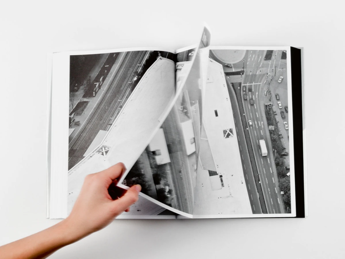 Photo book opened to a black-and-white aerial cityscape; a hand turns a page on a white surface.