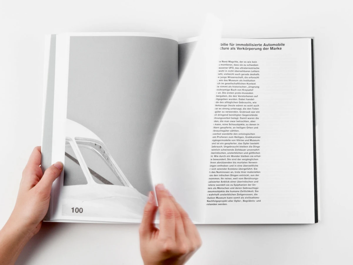 Hands turning an open magazine on a white surface; left page features a grayscale car image, right page shows dense text.