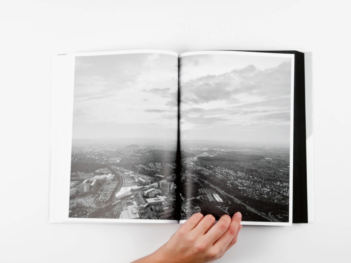Open book on white surface, displaying black-and-white aerial cityscape; a hand holds the bottom edge.