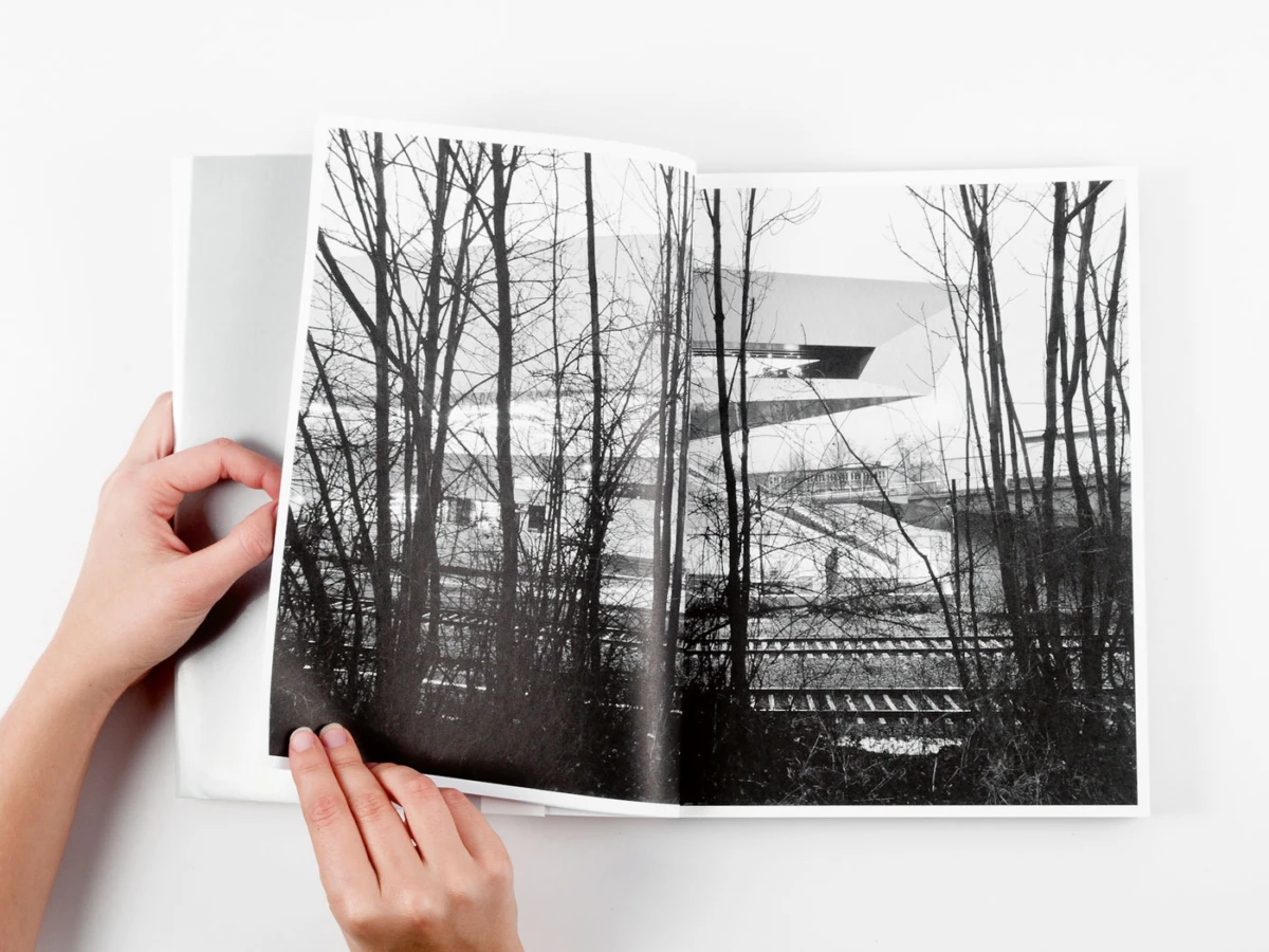 Open photo book held by hands, displaying a black-and-white landscape with leafless trees and railway tracks near a modern building.